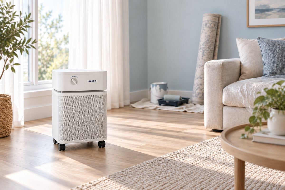White Austin Air HealthMate Plus air purifier on the floor of a bright, freshly redecorated living room with a paint can, roller tray, and a rolled-up new rug